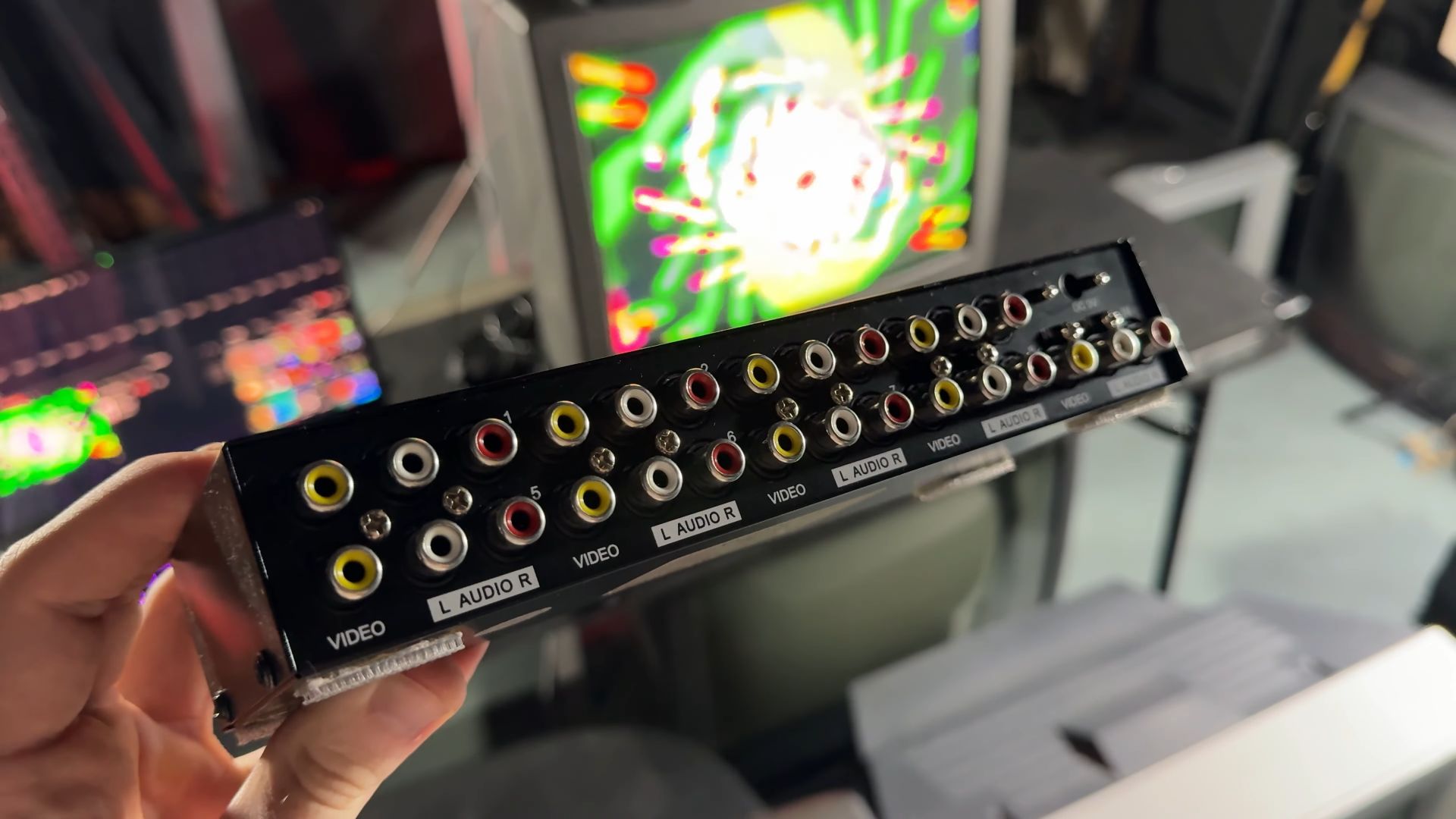 A hand holding a video splitter box with multiple RCA audio and video input/output ports, in front of a CRT television displaying colorful abstract graphics.