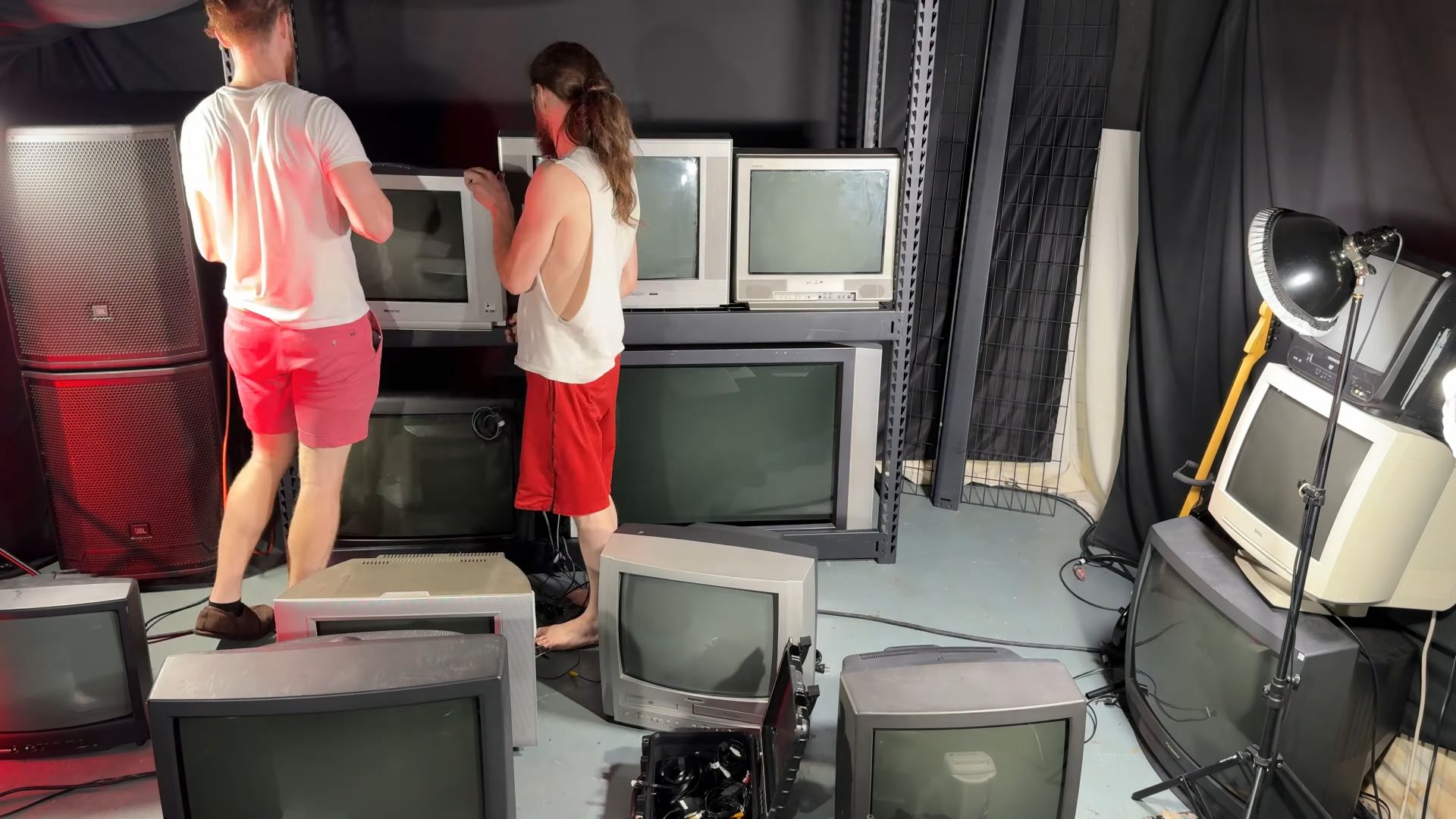 Tom and Ricky building a wall of CRT televisions using steel shelving, surrounded by various CRTs and studio lighting equipment.