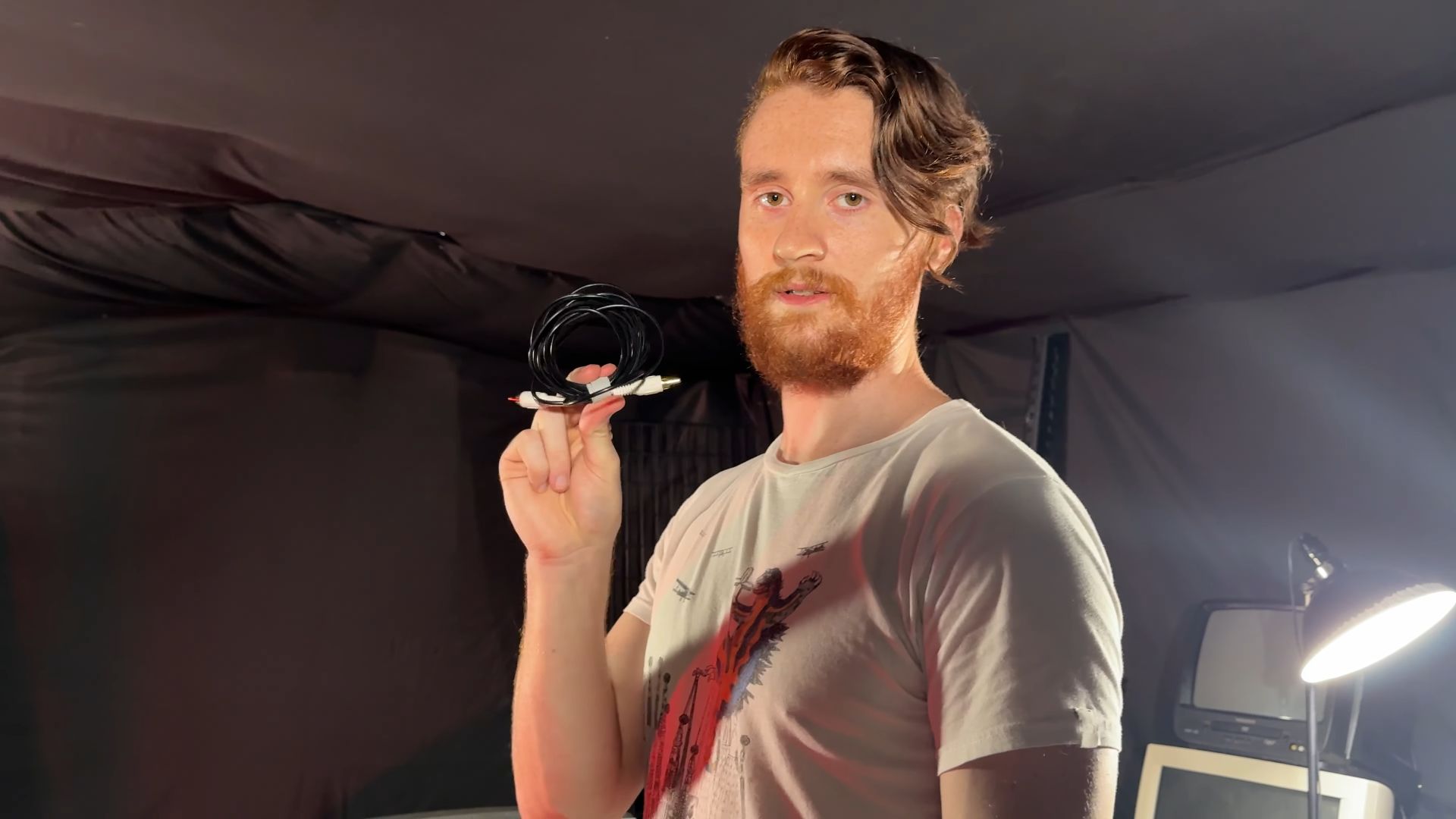 Tom holding a wrapped RCA cable, standing in a room with studio lighting and CRT televisions in the background.