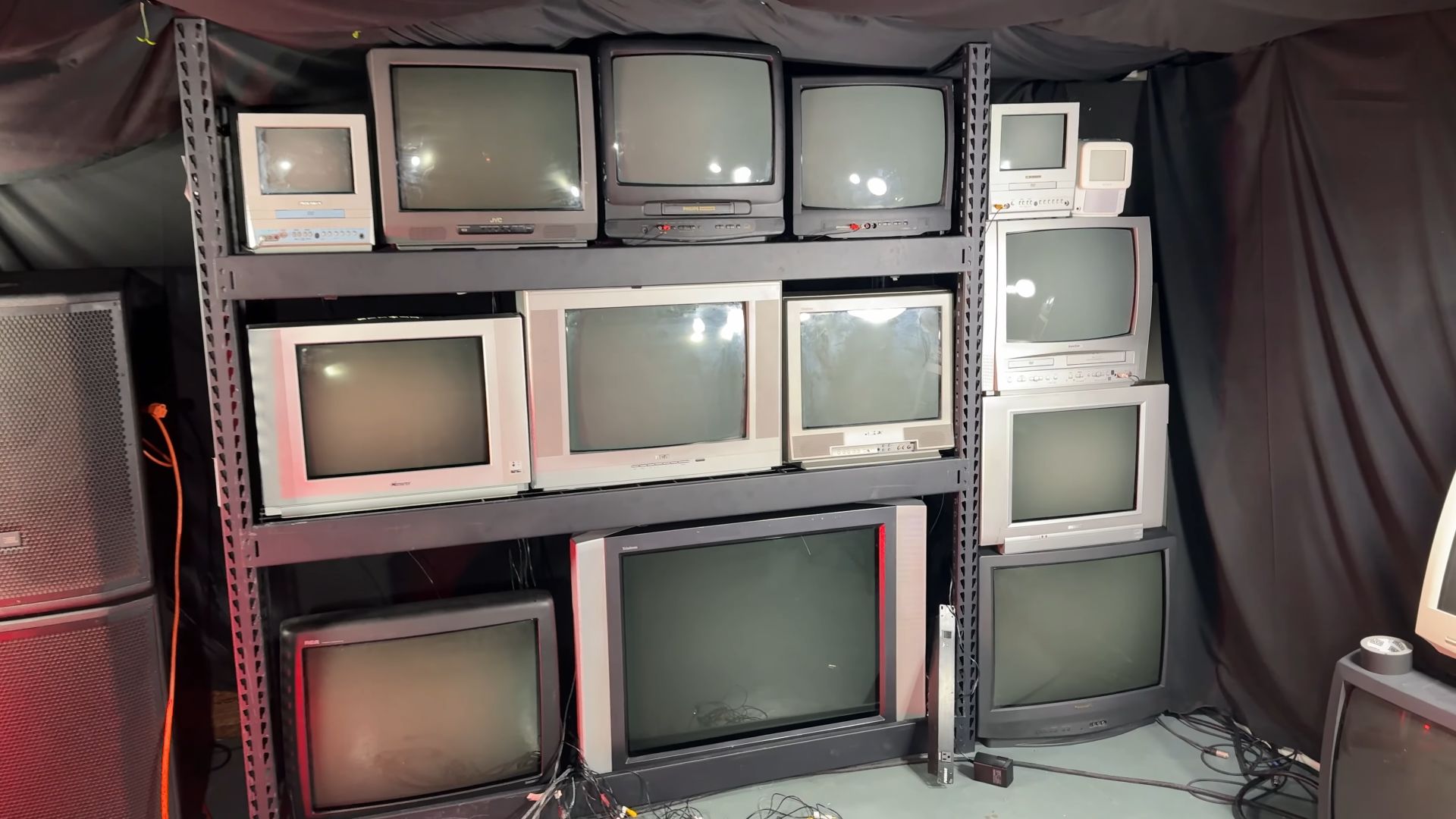 A completed wall of various CRT televisions arranged on steel shelving, surrounded by additional CRTs and audio equipment.