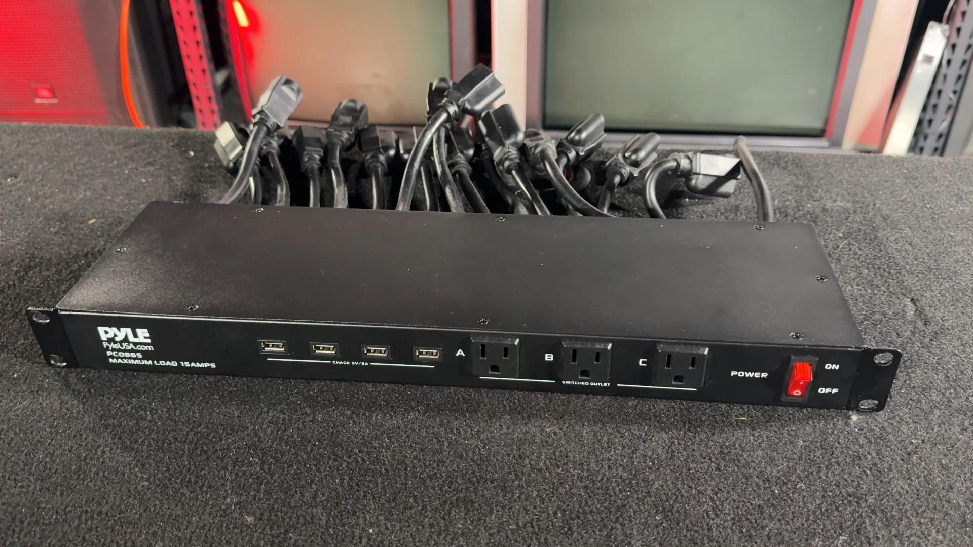 A Pyle power distribution unit (PDU) with multiple power cords plugged in, featuring three switched outlets labeled A, B, and C, and four USB ports for charging, with a maximum load of 15 amps.