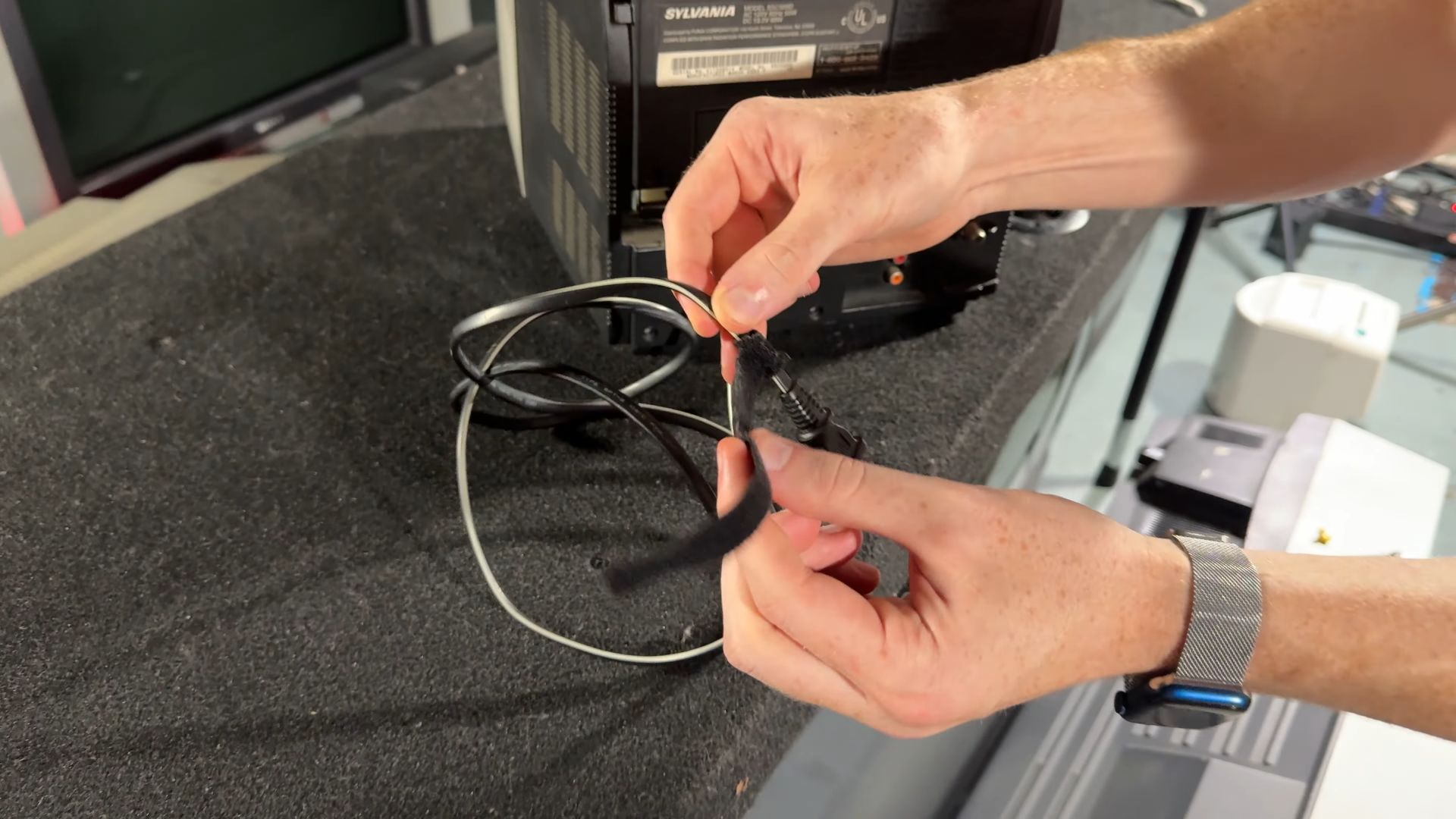 Tom is wrapping a CRT power cable using a cable wrap, with the back of a Sylvania CRT television visible in the background.