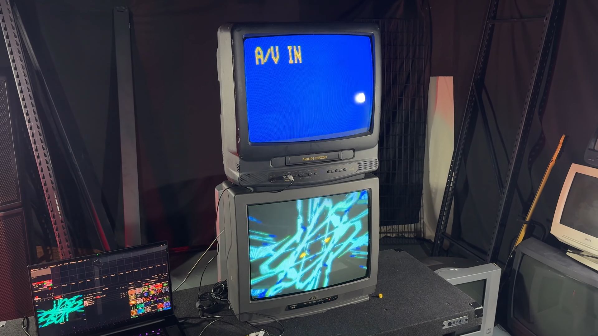 Two stacked CRT televisions, with the top Philips TV showing a blue screen with "A/V IN" text, and the bottom JVC TV displaying colorful abstract graphics. A laptop connected to the setup is on the left.