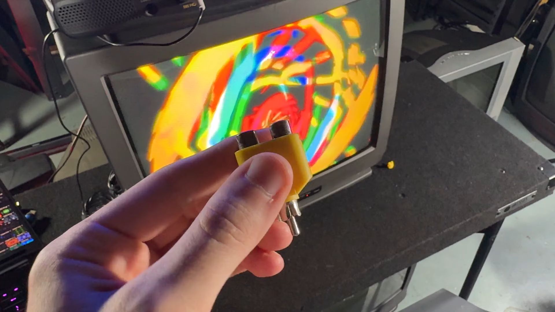 A hand holding a yellow RCA splitter in front of a vintage CRT television displaying colorful abstract graphics. The setup includes a laptop and another CRT television in the background.